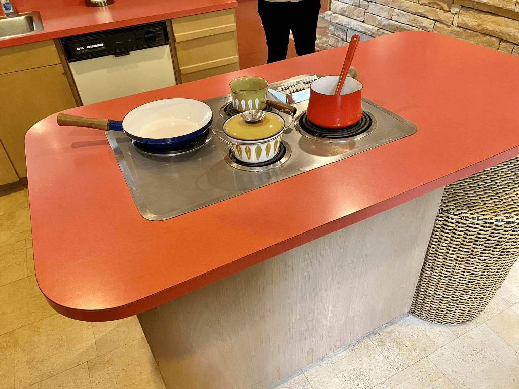 A red kitchen countertop featuring a frying pan, two small pots, and a saucepan on a stove, with a woven basket in the background.