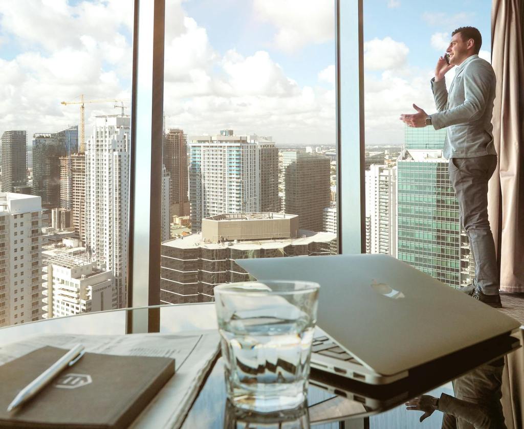 A professional man in a suit is standing by a large window in a high-rise building, talking on the phone while gesturing. In the foreground, a laptop and a glass of water are placed on the table.