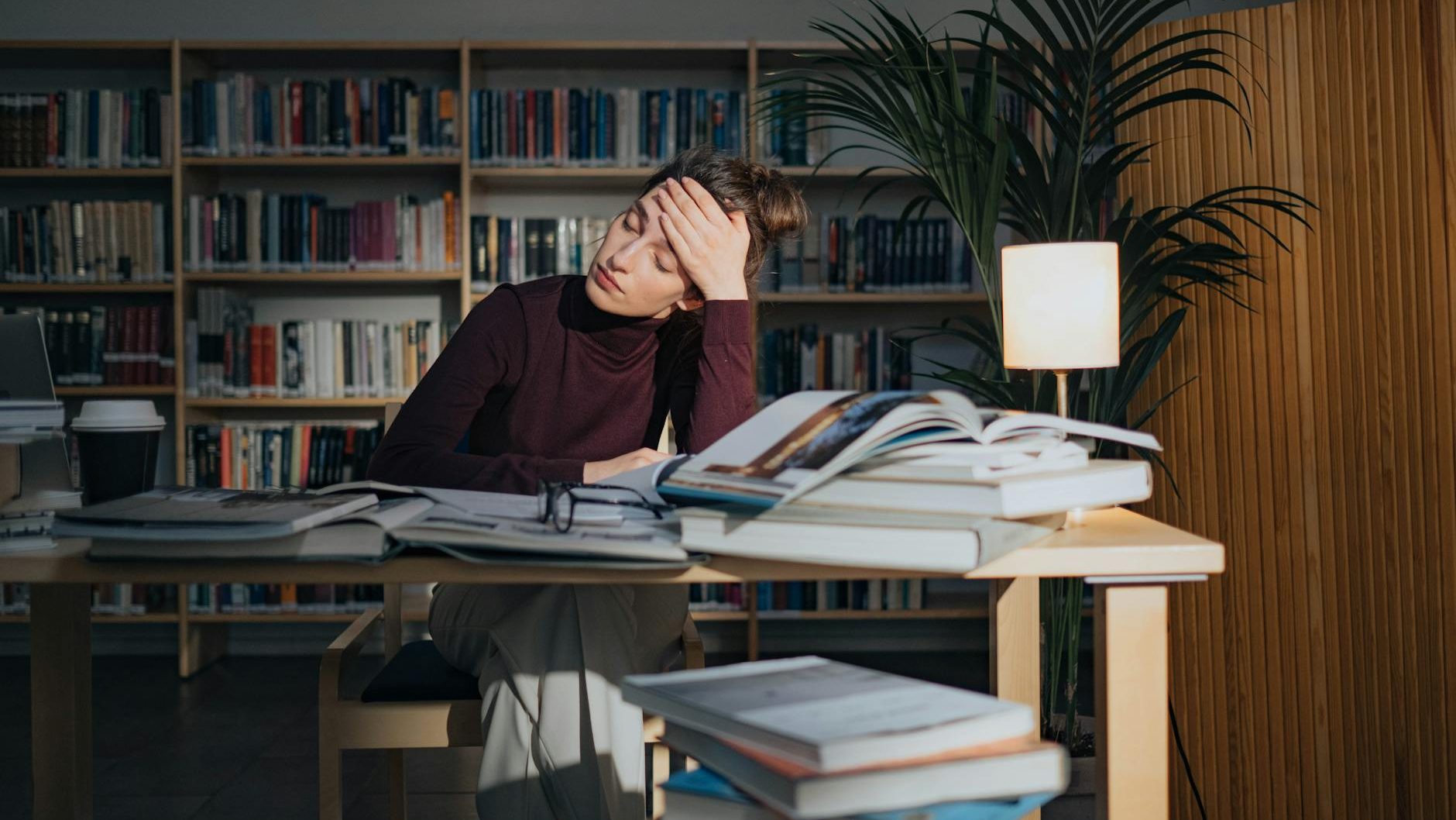 A young woman resting her head on her hand in a study environment, surrounded by stacks of books and a coffee cup, with a warm lamp casting light.
