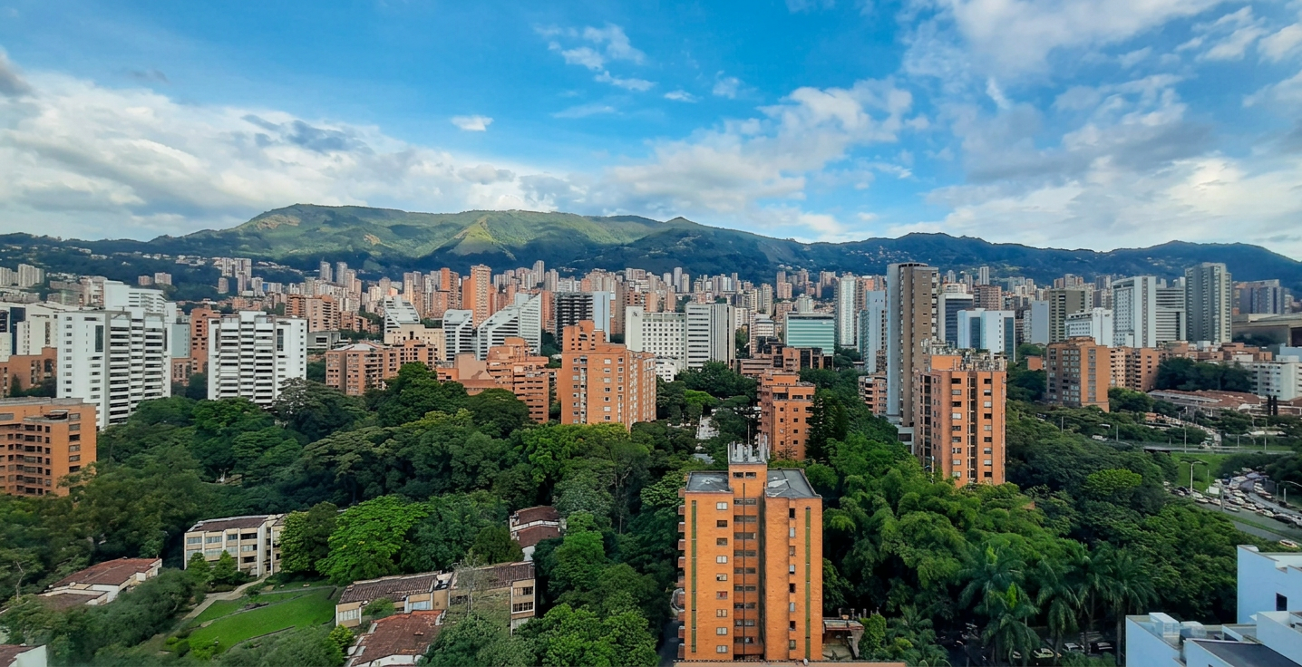 image A panoramic view of Medellín, Colombia, with numerous orange-brick and white high-rise apartment buildings surrounded by lush green trees. In the background, green mountains rise under a partly cloudy blue sky.