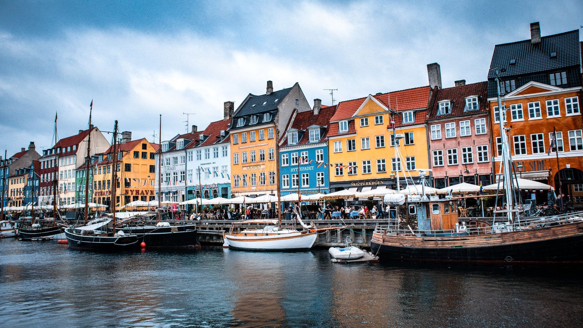 A row of colorful historic townhouses in yellow, blue, orange and red line a canal in Copenhagen's Nyhavn district. Traditional wooden sailboats are moored in the foreground, with outdoor cafe umbrellas and people sitting along the waterfront under a cloudy sky. The water reflects the buildings.